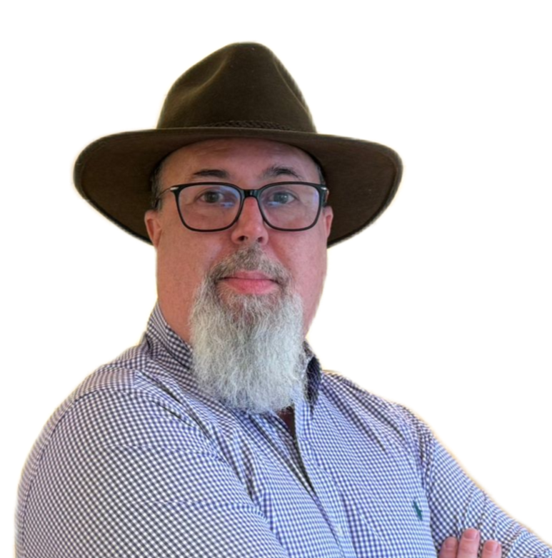 Trevor Gibbs is wearing a brown Fedora hat; he also has a long white/greyish beard, goatee style. He is wearing a purple shirt with small white squares on it and with his arms folded in front of him